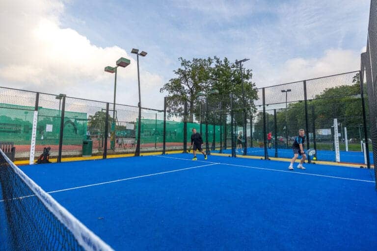 Men playing padel on an outdoor court at Oxshott Village Sports Club, with cloudy weather overhead and trees lining the background—capturing the club’s active atmosphere and scenic Surrey setting.