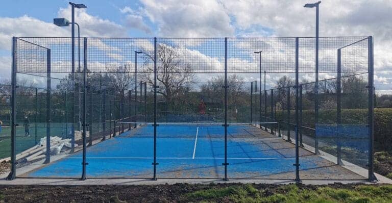 Empty padel court at Church Broughton Tennis Club, featuring enclosed glass walls, a net at center, and a clean synthetic surface, with no players present.