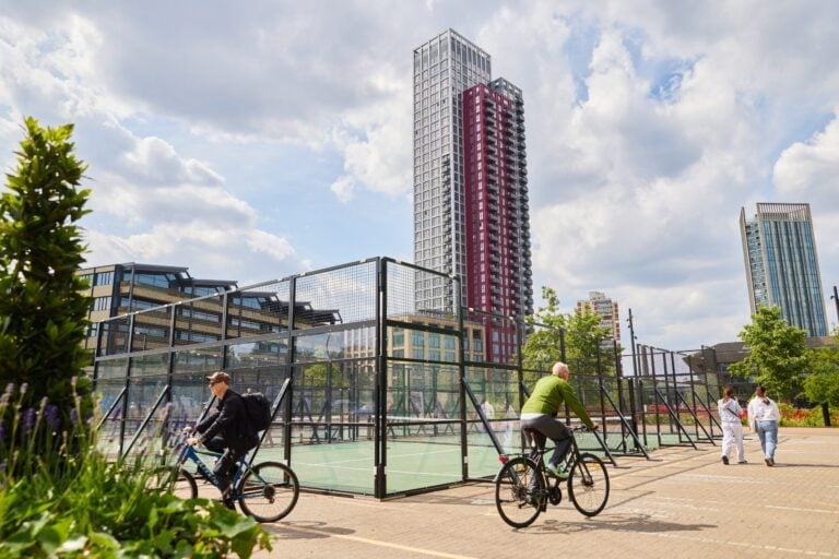 People cycling past open-air padel courts at Instant Courts, Canada Water, with a modern tower block in the background – urban padel tennis in London.