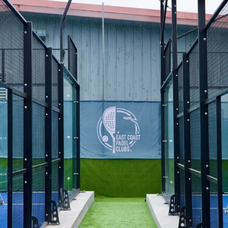 View down the middle of two panoramic padel courts at East Coast Padel Clubs, with the club logo prominently displayed, showcasing the modern outdoor facility and community-focused atmosphere.